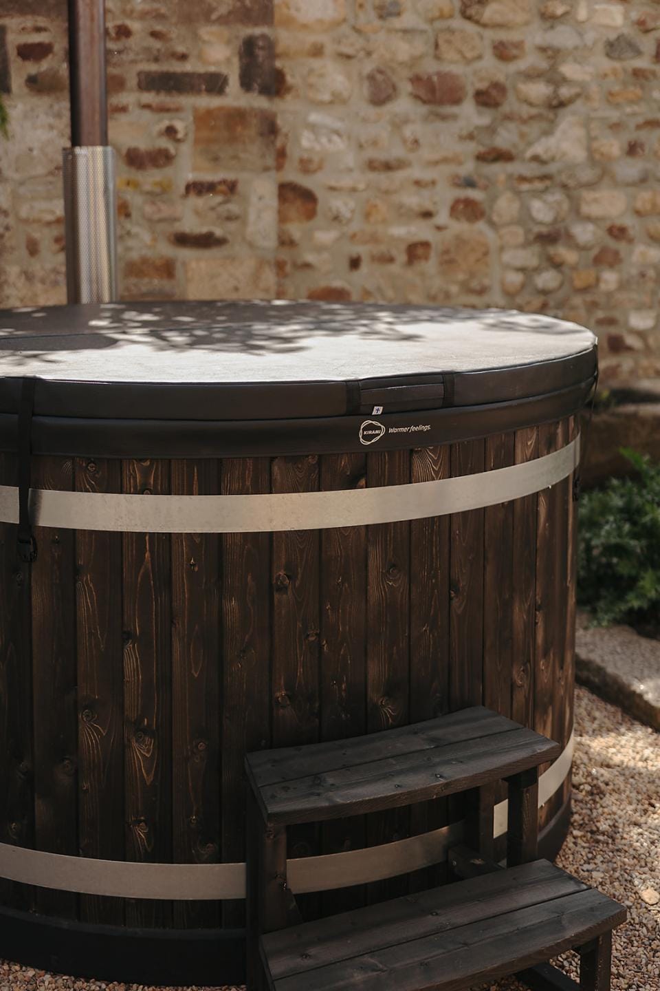 A close-up of a dark wood Kirami wood-fired hot tub fitted with a black insulating cover and matching two-tier wooden steps, set against a rustic stone wall on the Isle of Man.