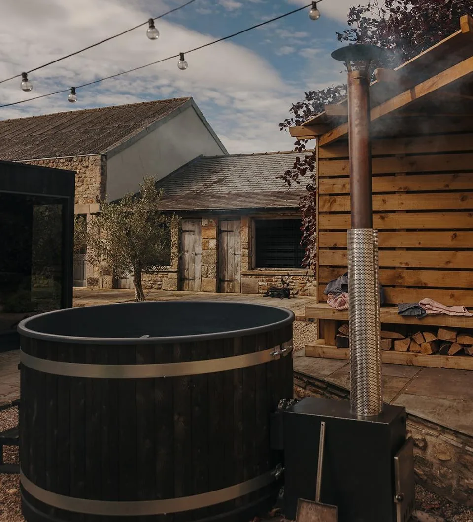 A dark wood Kirami wood-fired hot tub with a smoking chimney stove installed in a rustic courtyard next to a timber log store, festoon lights, and stone buildings on the Isle of Man.