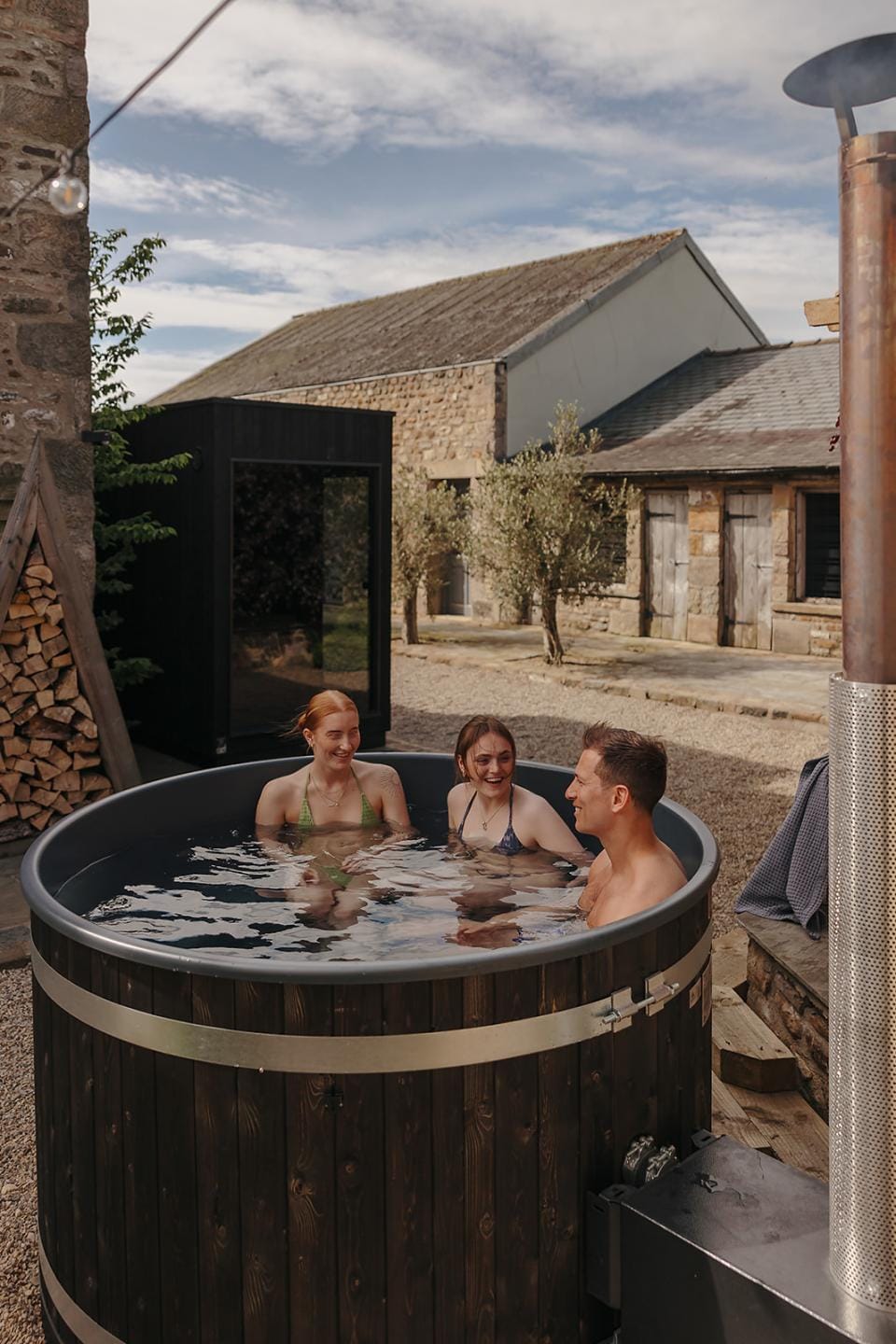 Three friends smiling and chatting inside a dark wood Kirami wood-fired hot tub, located in a rustic gravel courtyard with a modern black outdoor sauna building and old stone barns in the background on the Isle of Man.