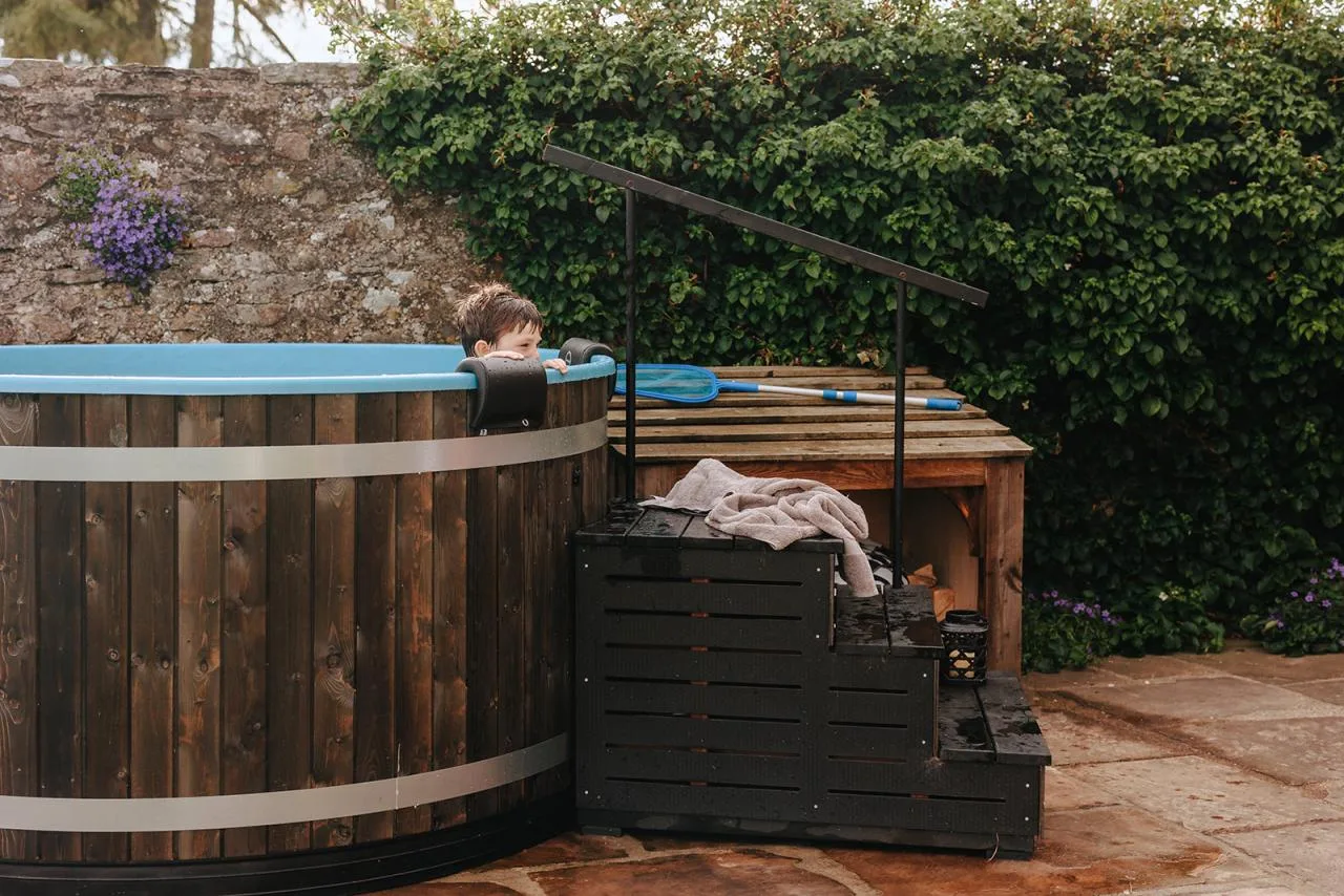 A young boy looking over the edge of a dark wood Kirami wood-fired hot tub with a blue liner, installed on a stone patio next to black three-tier safety steps with a matching metal handrail on the Isle of Man.
