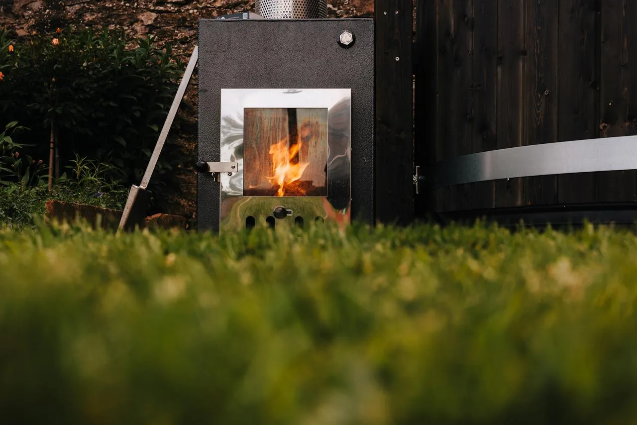 A low-angle shot taken from across a green lawn showing a bright orange fire crackling behind the polished glass window door of a black Kirami external hot tub heater on the Isle of Man.