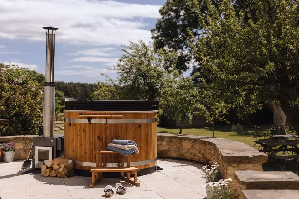 A round timber-clad Kirami wood-fired hot tub with an external log burner and tall chimney installed on a paved stone patio bounded by a low stone wall in an Isle of Man garden.