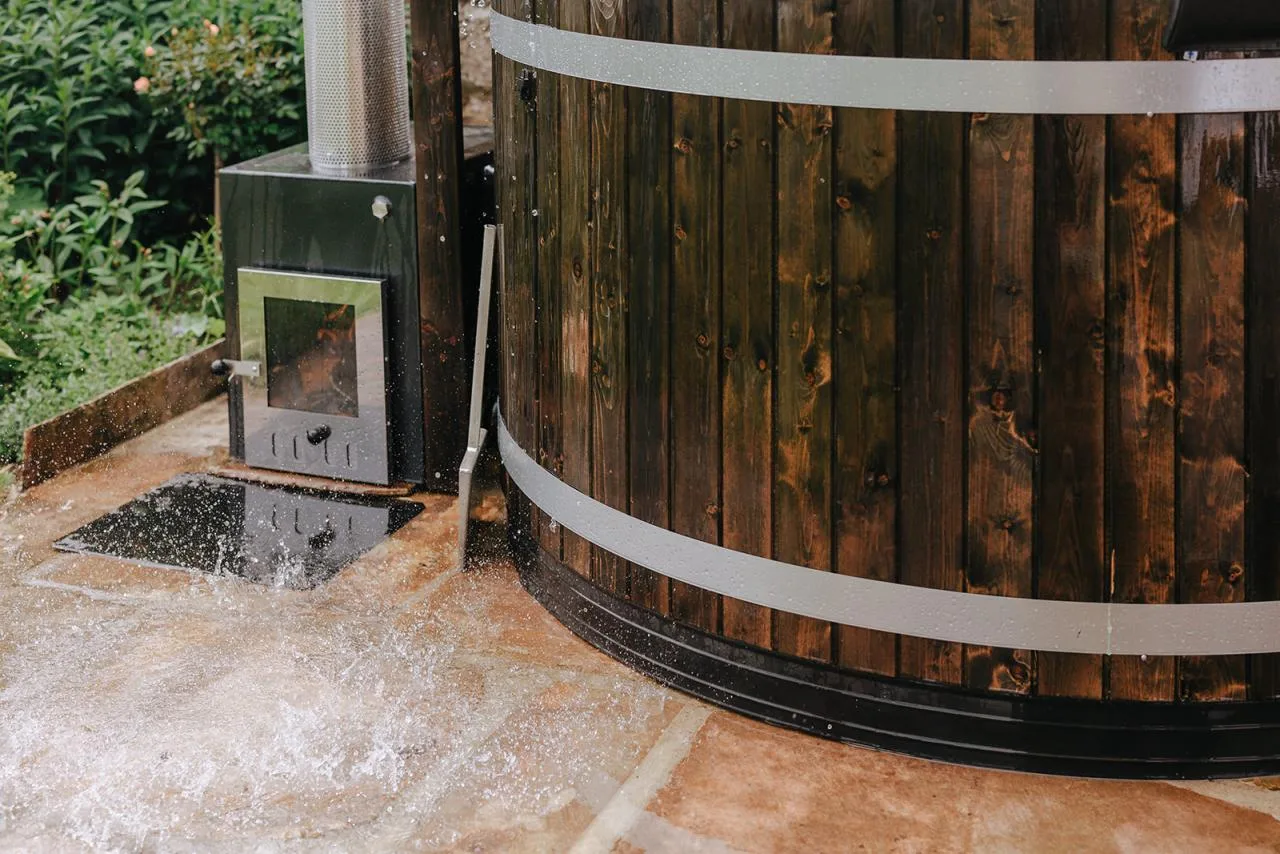 A close-up view of a dark wood Kirami Comfort Family L wood-fired hot tub on a flagstone patio, capturing water splashing onto the stone next to its external heater stove with a glass window door on the Isle of Man.