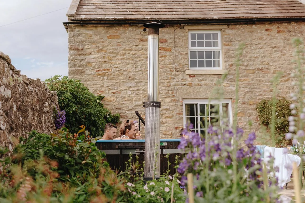A family relaxing inside a Kirami wood-fired hot tub in a scenic cottage backyard, framed by blooming purple flowers and a traditional stone house on the Isle of Man.