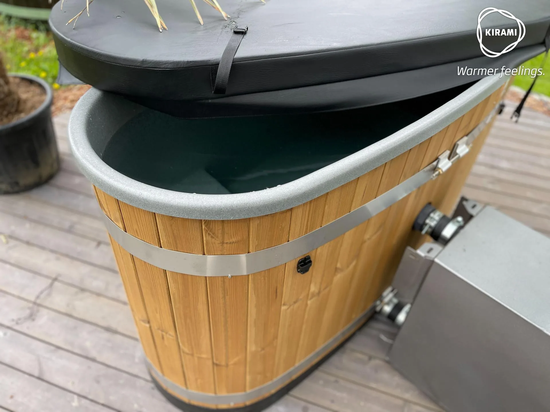 An oval Kirami Original Tiny wood-fired hot tub on a wooden deck, showing a thick black insulating cover partially lifted to reveal the water inside, located on the Isle of Man.
