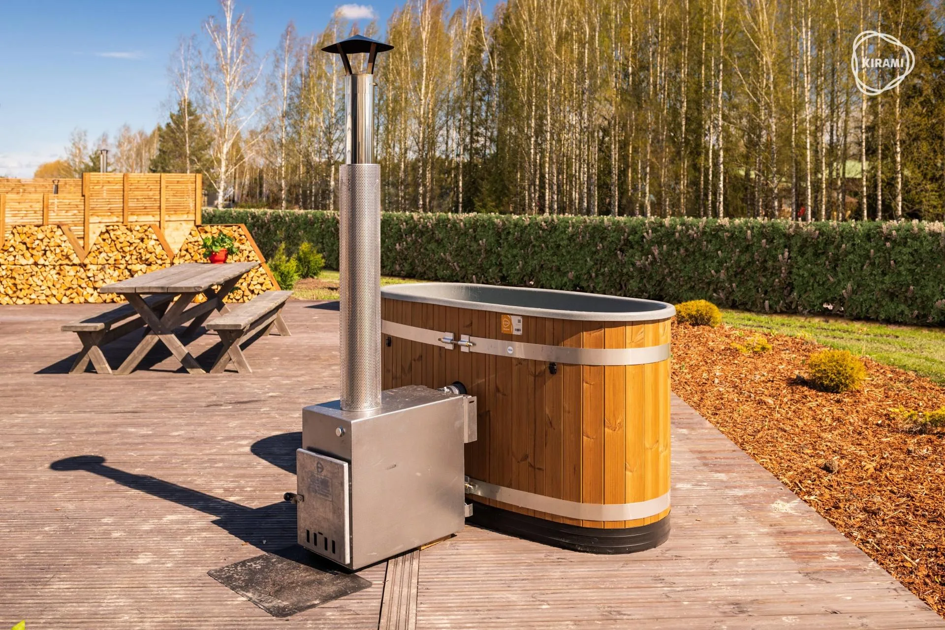 A Kirami Original Tiny wood-fired hot tub with an external stainless steel stove installed on a wide wooden deck next to a picnic table and a log store wall on the Isle of Man.