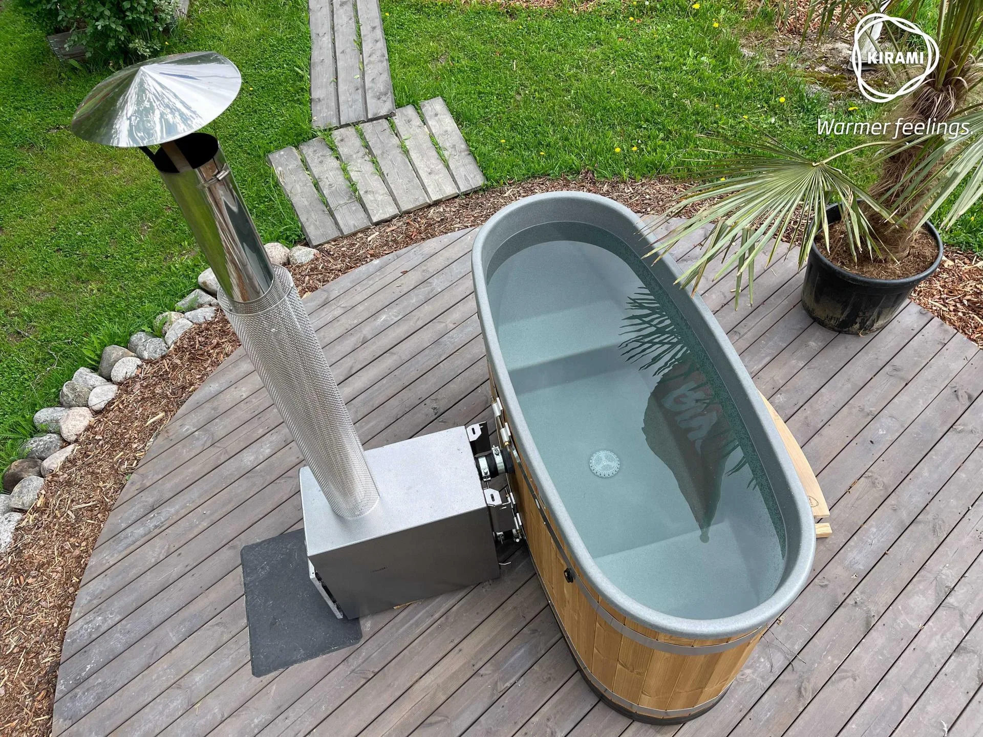 An overhead view of a compact oval Kirami Original Tiny wood-fired hot tub with an external heater and tall chimney stove, installed on a rounded wooden deck in an Isle of Man garden.