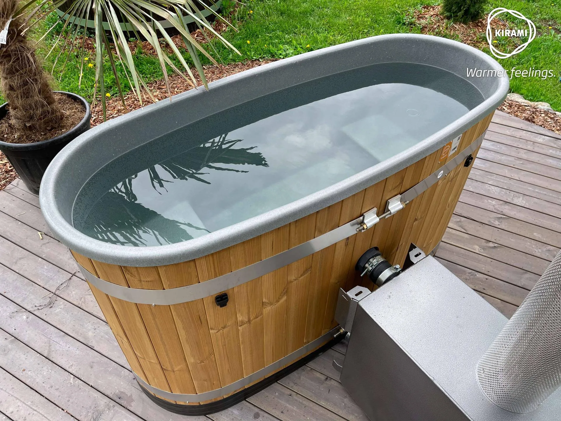 An angled side view of a filled Kirami Original Tiny wood-fired hot tub on a timber deck, showing the pipe connections leading to its stainless steel external wood burner on the Isle of Man.