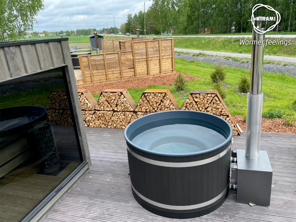 A black Kirami wood-fired hot tub filled with water on a large timber deck next to a modern outdoor sauna building, featuring stacked triangular log stores in the background on the Isle of Man.