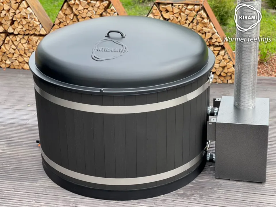A black Kirami wood-fired hot tub fitted with a matching hard plastic black dome cover and connected to an external stove heater, sitting on a wooden deck on the Isle of Man.