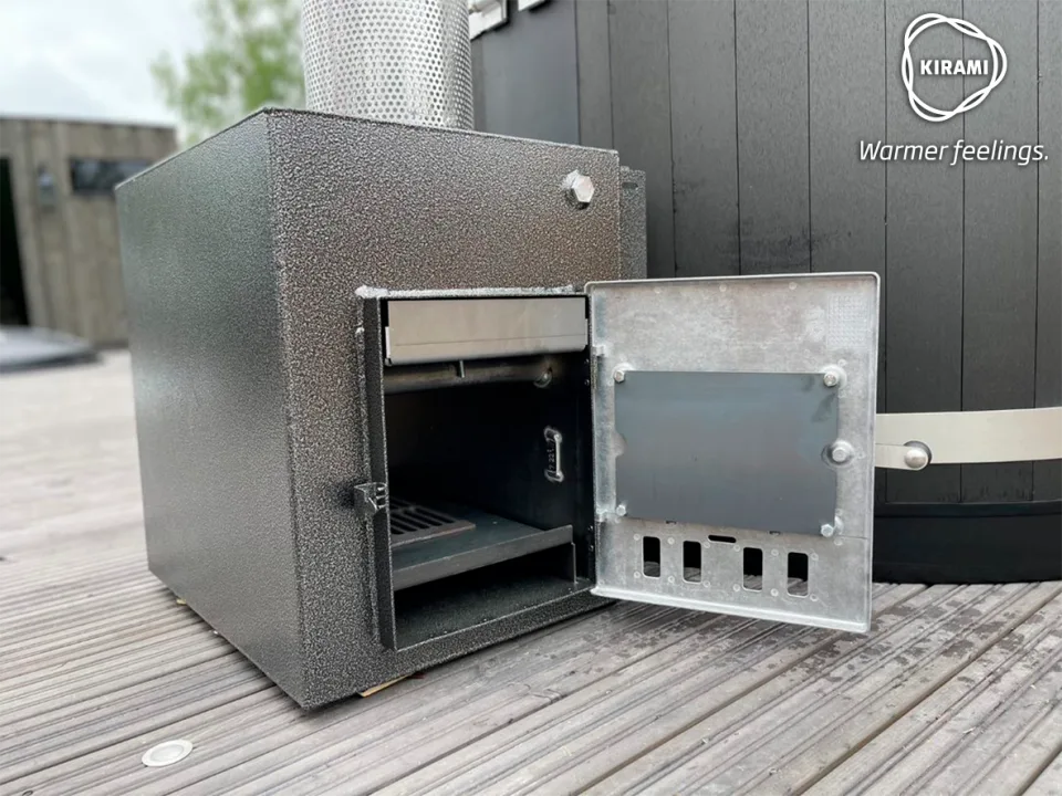 A close-up view of a grey textured Kirami external wood-burning stove with its metal door open, revealing the firebox chamber next to a black hot tub on a wooden deck on the Isle of Man.
