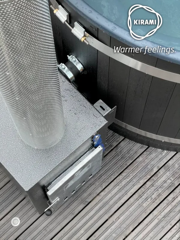 A top-down close-up shot of a black Kirami wood-fired hot tub showing the pipe connection and tension bands leading to the stainless steel external heater stove on a wooden deck on the Isle of Man.