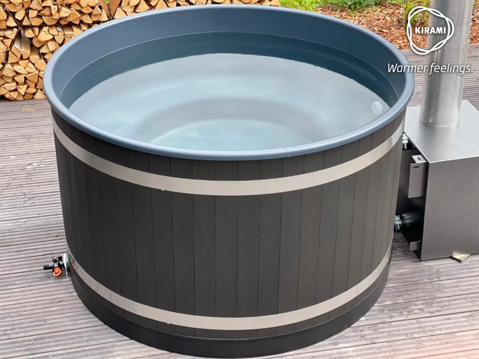 A side view of a round Kirami wood-fired hot tub with a dark grey exterior and smooth inner liner, filled to the brim with clear water and sitting on a timber patio deck on the Isle of Man.