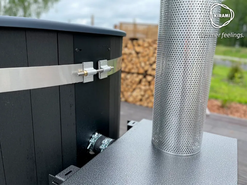 A close-up view focusing on the perforated stainless steel chimney heat shield and secure side plumbing connections of a black Kirami wood-fired hot tub on the Isle of Man.