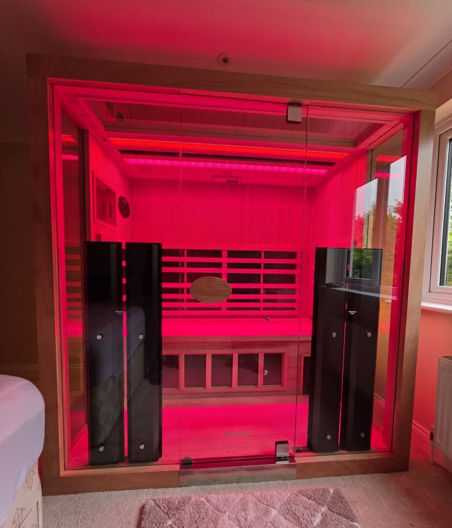 A direct front view of a large 3-person Jacuzzi infrared sauna installed in a residential room, completely illuminated by vibrant red light therapy with a full glass facade.