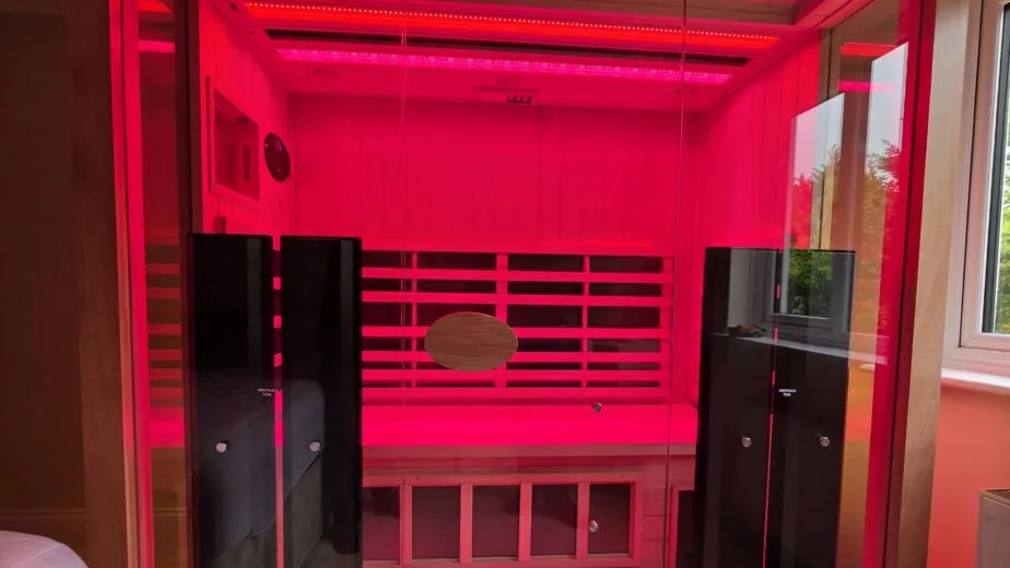 A direct front view of a large 3-person Jacuzzi infrared sauna installed in a residential room, completely illuminated by vibrant red light therapy with a full glass facade.