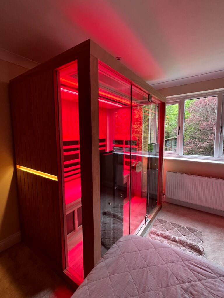 A left three-quarter angled view of a large 3-person Jacuzzi infrared sauna in a residential bedroom, featuring a glowing amber exterior light strip and intense red light therapy illuminating the interior.