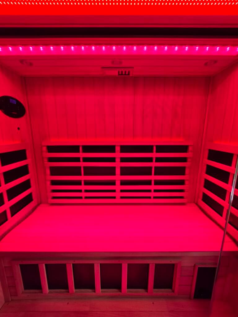 A straight-on interior view of a 3-person Jacuzzi infrared sauna bench completely bathed in intense red light therapy, featuring slatted wood backrests and an oval wall control panel.