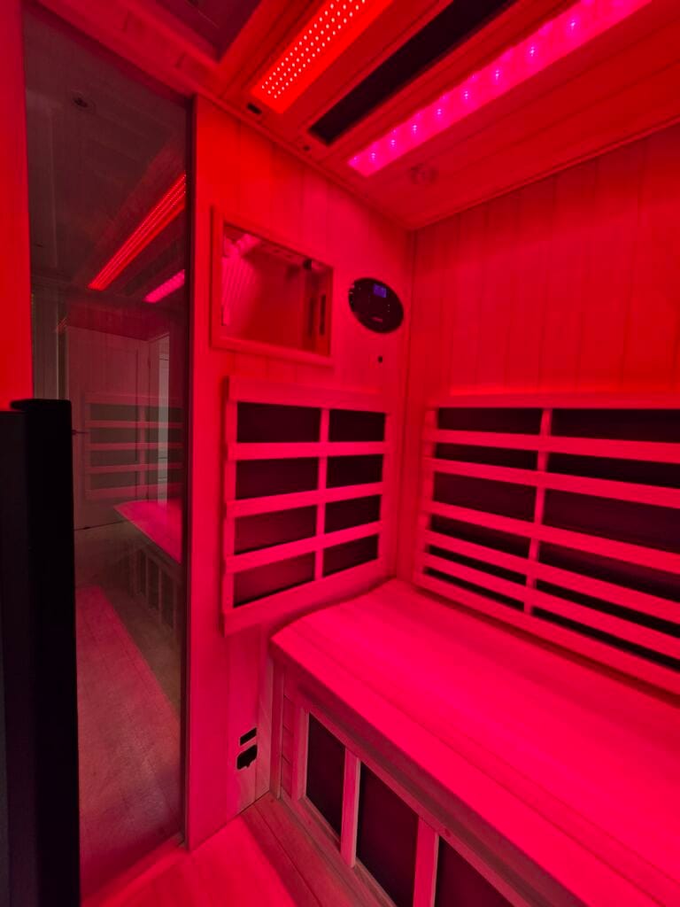 An interior view of a 3-person Jacuzzi infrared sauna completely illuminated by red light therapy ceiling panels, showcasing the timber benches, slatted backrests, and a wall control panel.