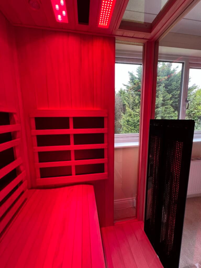An interior view of a Jacuzzi infrared sauna glowing under intense red light therapy, looking past a tall black heater panel and glass partition toward a window showing evergreen trees.