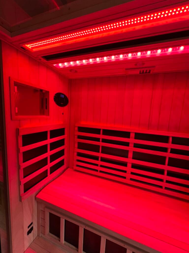 An interior corner view of a Jacuzzi infrared sauna bathed in vibrant red light from overhead therapy strips, highlighting the cedar benches and slatted wooden backrests.