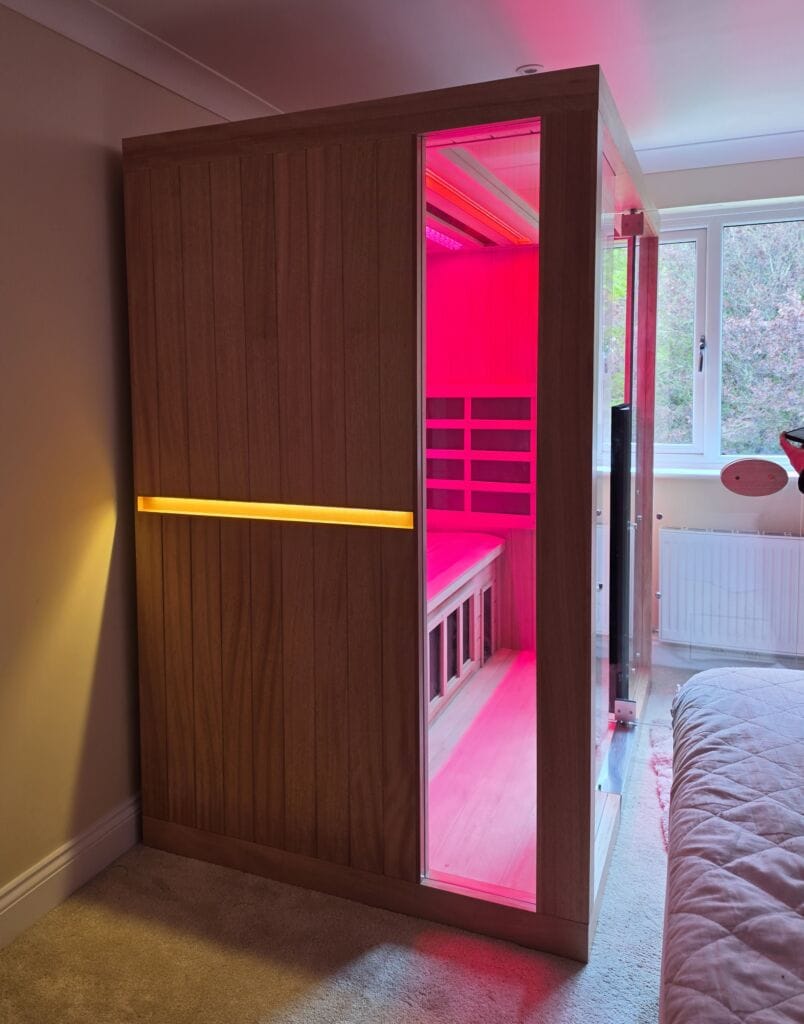 A three-quarter side view of a wooden Jacuzzi infrared sauna installed in a residential bedroom, featuring a glowing amber exterior light strip and a vibrant pink chromotherapy interior.