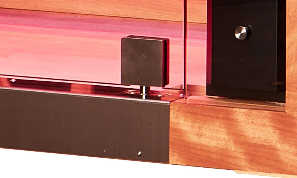 A detailed close-up shot of the robust lower metal hinge, glass door threshold, and rich wood framing of a Jacuzzi infrared sauna illuminated with a soft pink internal chromotherapy glow.