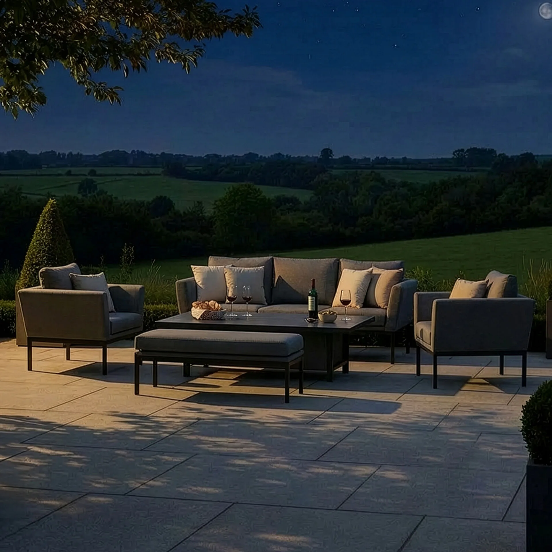 The Pulse zinc grey outdoor fabric garden sofa set arranged on a patio illuminated by warm evening lighting under a dark night sky in the Isle of Man.
