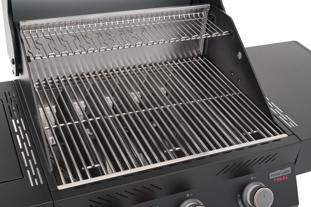 A detailed interior view of a Primeliner gas grill, featuring heavy-duty stainless steel cooking grates and an upper warming rack, with the "Prime Liner Ebull" branding visible on the side panel.
