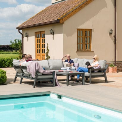 A group of friends relaxing on the contemporary horseshoe shape Maze New York garden sofa set, featuring thick showerproof cushions and a slatted aluminium coffee table by a pool.