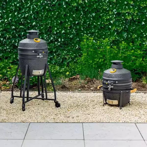 Two matte black GRLLR Ember Classic 16-inch ceramic kamado grills side by side on a gravel patio area—one mounted on a tall mobile trolley and the other in a compact tabletop nest.