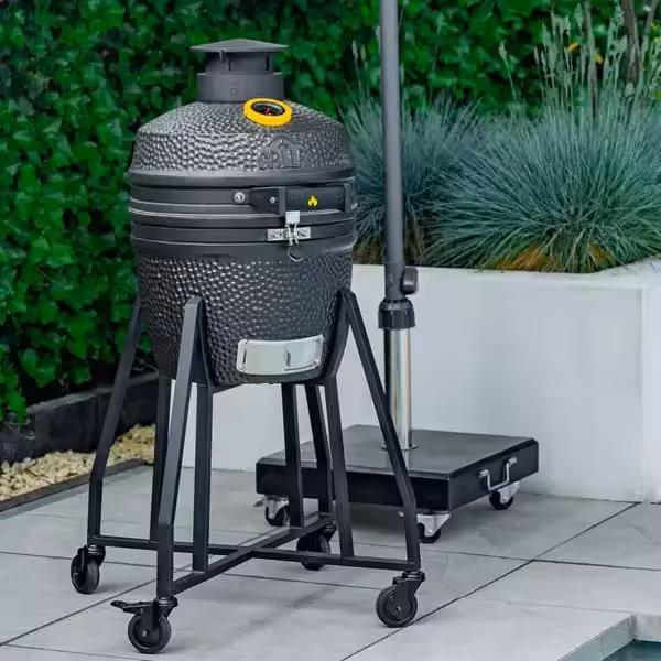 A compact matte black GRLLR Ember Classic 16-inch ceramic kamado grill on a mobile trolley set on a grey paved patio next to an outdoor umbrella stand and garden planter.