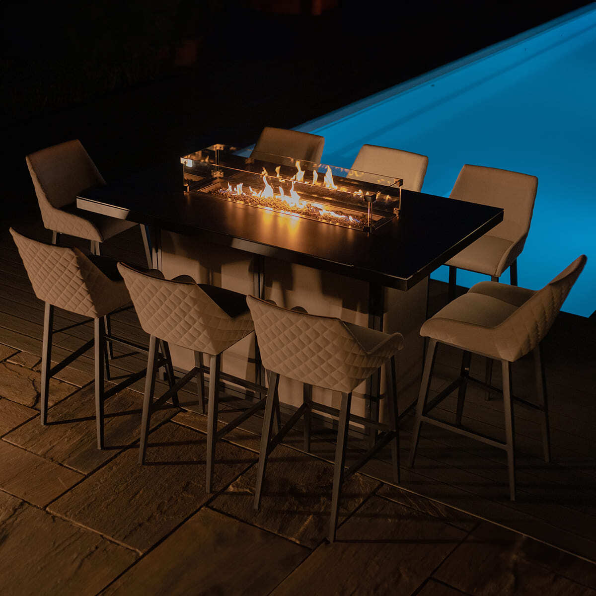 The Maze - Outdoor Fabric Regal 8 Seat Rectangular Bar Set Latte with café frame illuminated by its integrated gas firepit at night next to a lit swimming pool at Roberts Leisure, Isle of Man.