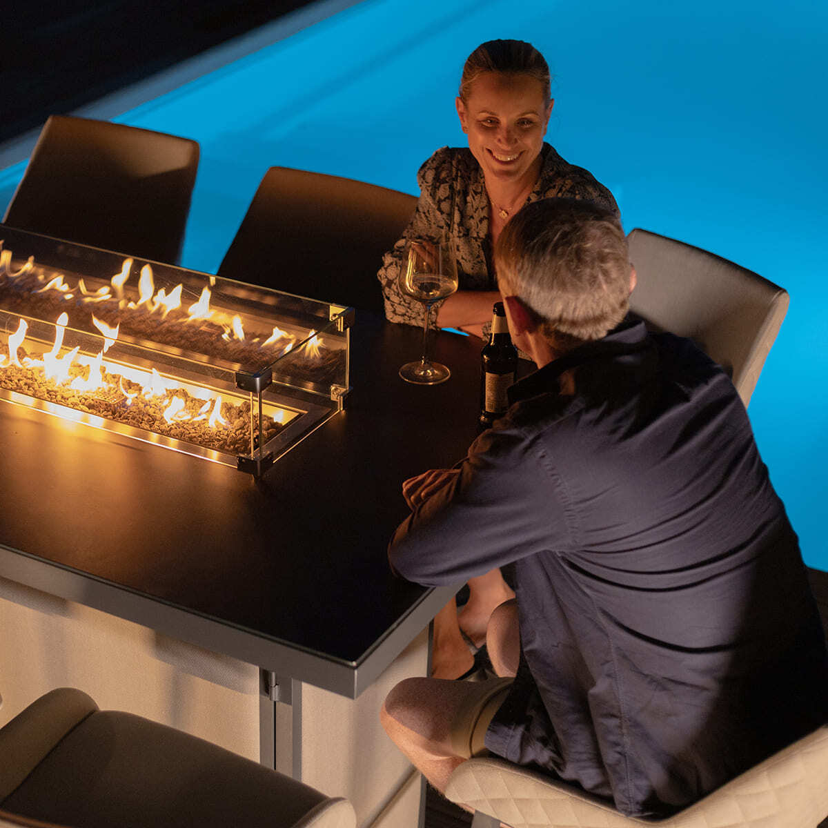 A couple enjoying drinks around the lit firepit of the Maze - Outdoor Fabric Regal 8 Seat Rectangular Bar Set Latte with café frame at night, from Roberts Leisure, Isle of Man.