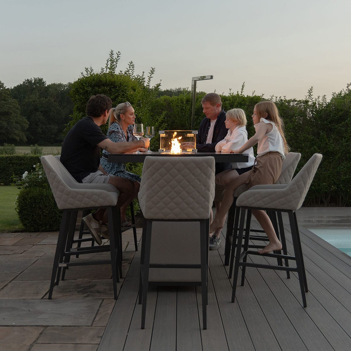 A family gathered around the lit firepit of the Maze - Outdoor Fabric Regal 8 Seat Rectangular Bar Set Latte with café frame on a sunny evening at Roberts Leisure, Isle of Man.