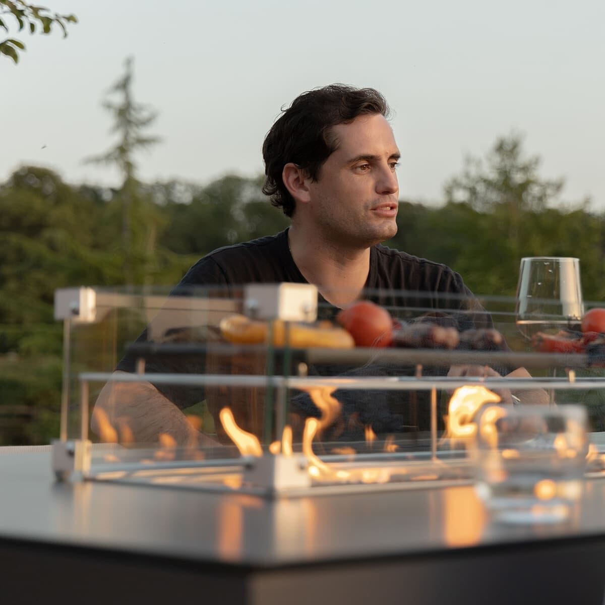 A man relaxing by the integrated gas firepit of the Maze - Outdoor Fabric Regal 8 Seat Rectangular Bar Set Latte with café frame, showcasing the protective glass surround at Roberts Leisure, Isle of Man.